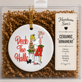 A round white ceramic Christmas ornament in clear retail packaging, featuring a retro-style illustration of a woman decorating a tree with the words 'Deck The Halls' in red. A hanging string is attached, and a label on the package reads 'Handsome Sam's' and 'Ceramic Ornament.