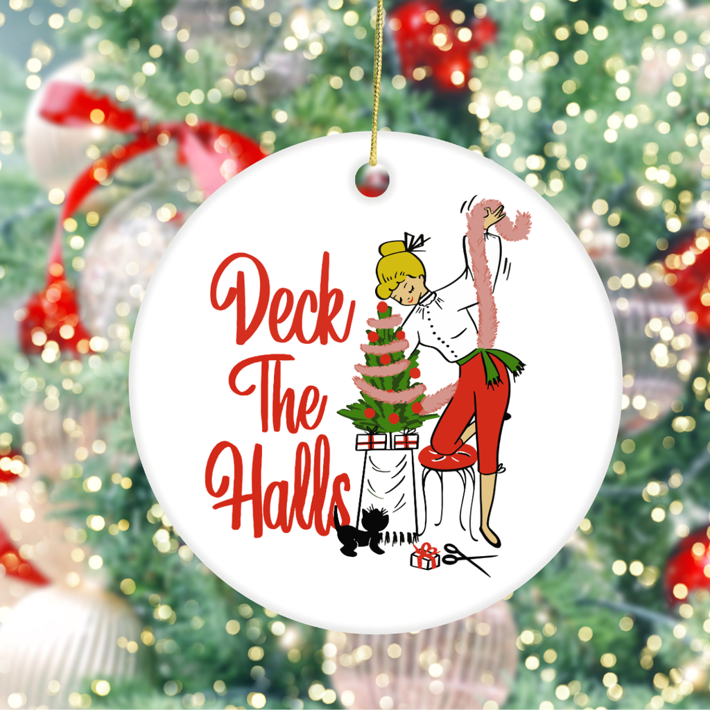 A round white ceramic Christmas ornament hanging from a gold string, featuring retro-style red text reading 'Deck the Halls' and an illustration of a woman decorating a Christmas tree. The ornament is photographed in front of a blurred Christmas tree with lights and ornaments.